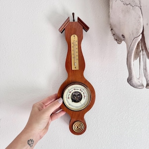 May include: A wall-mounted wooden weather station featuring a vertical thermometer with Fahrenheit markings and a circular barometer dial with text reading Rising, Rain, Change, Fair, and Very Dry, finished with a brass-colored circular accent.