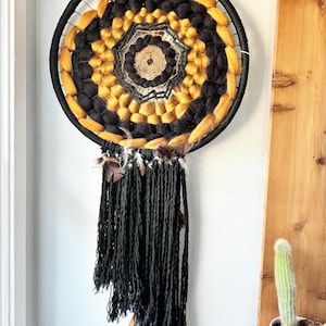 May include: A handcrafted dreamcatcher with a black, gold, and tan woven design. The circular frame is wrapped in black cord, with a central woven pattern and hanging black yarn tassels. Feathers are attached at the bottom.