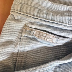 May include: Close-up of light blue denim jeans. The jeans feature a pocket with a small white tag that reads "Wrangler" in a cursive font. The fabric has a textured appearance, and the stitching is visible along the seams.