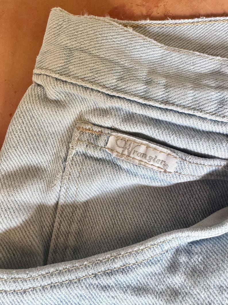 May include: Close-up of light blue denim jeans. The jeans feature a pocket with a small white tag that reads "Wrangler" in a cursive font. The fabric has a textured appearance, and the stitching is visible along the seams.
