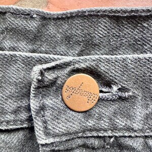 Op de afbeelding: Close-up van grijze denimstof met een koperen knoop. De knoop is rond en heeft het woord "seafarer" erin gegraveerd. De denim heeft een getextureerde uitstraling.
