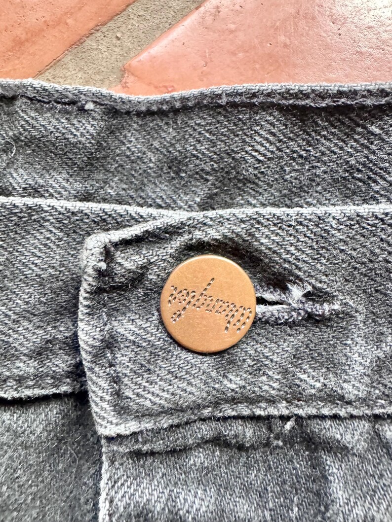 Op de afbeelding: Close-up van grijze denimstof met een koperen knoop. De knoop is rond en heeft het woord "seafarer" erin gegraveerd. De denim heeft een getextureerde uitstraling.