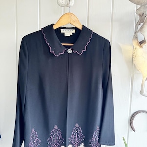 May include: Black blazer with a scalloped collar and hem. The collar and hem are trimmed with purple embroidery. The blazer has a button closure and long sleeves.