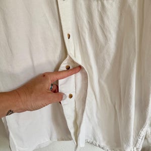 May include: Close-up of a white button-down shirt with a crochet pocket. The shirt has brown buttons and a textured, woven pocket. The fabric appears to be a soft, light material.