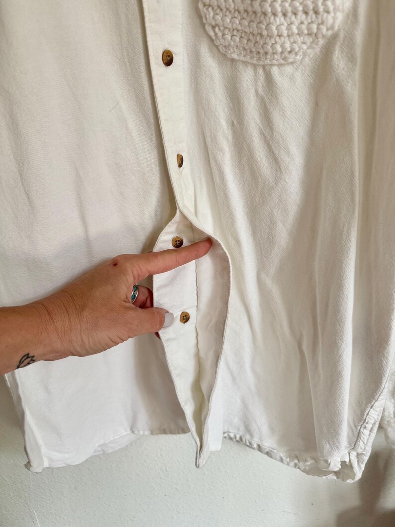 May include: Close-up of a white button-down shirt with a crochet pocket. The shirt has brown buttons and a textured, woven pocket. The fabric appears to be a soft, light material.