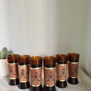 May include: Set of six vintage amber-colored highball glasses. Each glass features a wooden band with a skull and crossed swords design, accented with gold-toned metal trim. The glasses have a flared base.