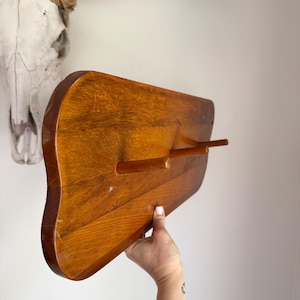 May include: A wooden wall-mounted coat rack with three hooks. The rack is made of light brown wood and has a slightly curved shape.