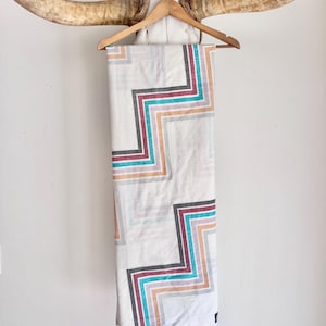 May include: A rectangular white textile featuring a repeating geometric pattern of stepped, zigzag lines in shades of dark gray, deep red, bright turquoise, and soft peach. The fabric hangs vertically from a wooden hanger.