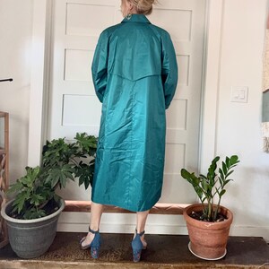May include: Teal-colored long coat with a collared neckline and long sleeves. The coat is knee-length and has a slight sheen. The person is wearing blue heels. The background includes a white door, potted plants, and a wooden surface.