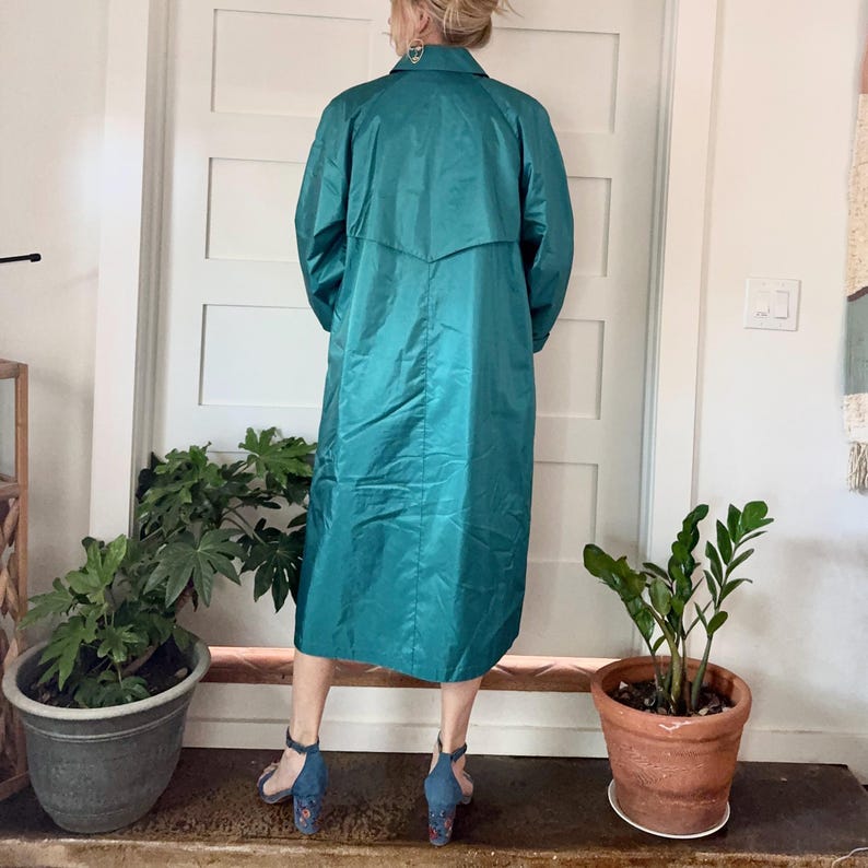 May include: Teal-colored long coat with a collared neckline and long sleeves. The coat is knee-length and has a slight sheen. The person is wearing blue heels. The background includes a white door, potted plants, and a wooden surface.
