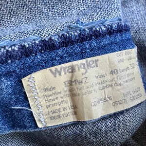 May include: Close-up of blue denim jeans with a Wrangler label. The label shows a waist size of 101.6 cm and a length of 81.3 cm. The jeans are made of 100% cotton. The denim has a textured appearance.