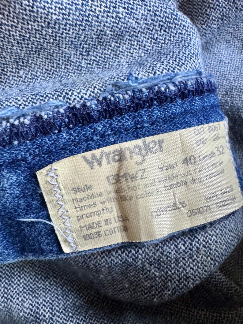 May include: Close-up of blue denim jeans with a Wrangler label. The label shows a waist size of 101.6 cm and a length of 81.3 cm. The jeans are made of 100% cotton. The denim has a textured appearance.