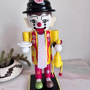 May include: A wooden clown nutcracker with a black hat and a yellow jacket over a red and white polka dot vest. The clown holds a white plate and a yellow bird-shaped toy. The base is black and yellow.