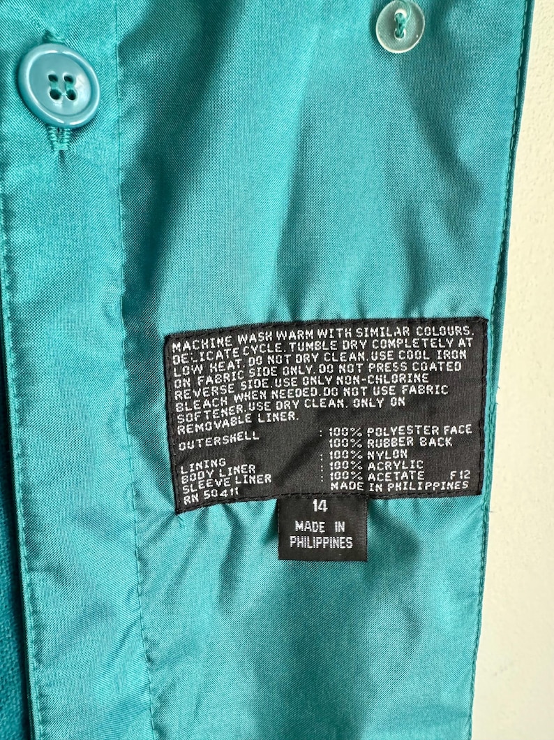 May include: Close-up of a teal jacket interior with a label indicating size 14 and "Made in Philippines." The label also lists fabric composition and care instructions. Teal buttons are visible.