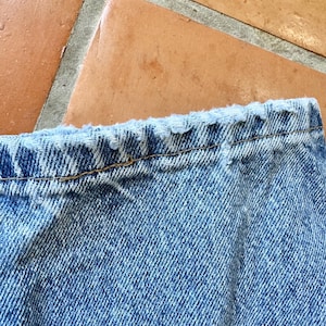 May include: Close-up of the top edge of a pair of light blue denim jeans. The jeans have a distressed, frayed edge along the waistband. The denim fabric shows a classic twill weave.