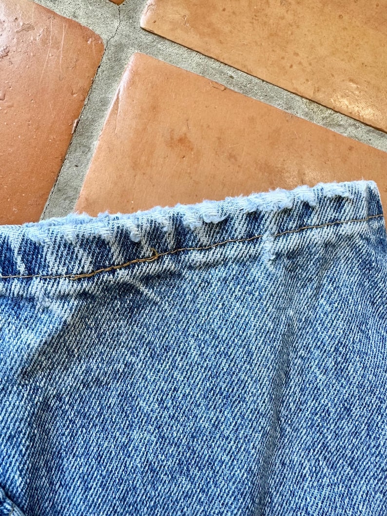 May include: Close-up of the top edge of a pair of light blue denim jeans. The jeans have a distressed, frayed edge along the waistband. The denim fabric shows a classic twill weave.