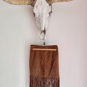 May include: A brown suede skirt featuring a straight-cut silhouette and a long fringe hemline. The skirt is suspended from a wooden hanger, showcasing a soft, matte texture and consistent brown coloring throughout the garment's fabric.
