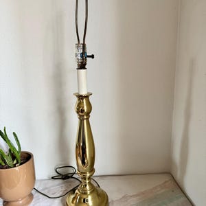 May include: A vintage brass table lamp with a classic candlestick design. The lamp features a polished gold finish, a white bulb socket, and a black on/off switch. The lamp is approximately 20 inches tall.