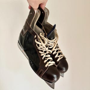 May include: A pair of vintage ice skates with brown leather uppers and white laces. The skates have a black accent on the sides and silver blades. The skates are held up against a white background.