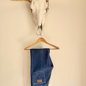 May include: Blue denim jeans hanging on a wooden hanger. The jeans have a classic five-pocket design with a brown leather patch on the back pocket. Above the jeans is a cow skull with long horns.