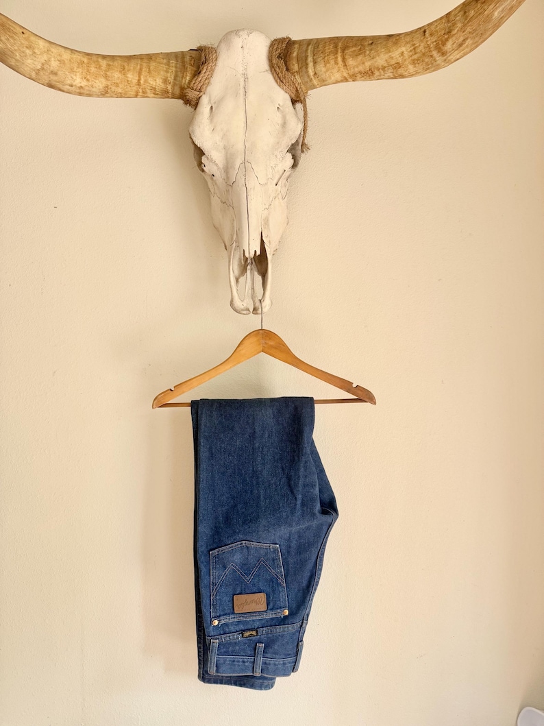 May include: Blue denim jeans hanging on a wooden hanger. The jeans have a classic five-pocket design with a brown leather patch on the back pocket. Above the jeans is a cow skull with long horns.