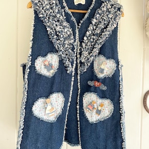 May include: A blue denim vest with a white and blue ruffled trim around the neckline. The vest has five heart-shaped patches with embroidered floral patterns and appliqued figures of children and animals.