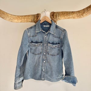 May include: Light blue denim shirt with a classic collar, two chest pockets, and button-down closure. The sleeves are cuffed, and the shirt is hanging on a wooden hanger. The shirt is displayed against a neutral background.