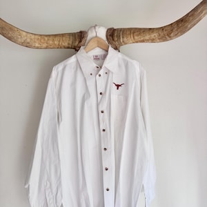 May include: A crisp white long-sleeve button-down shirt featuring a small, embroidered dark red longhorn steer logo on the left chest pocket area. The shirt has a classic collar, a full front button placket, and buttoned cuffs on the sleeves.