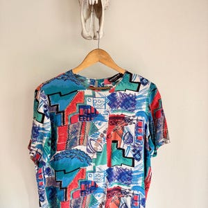 May include: A short-sleeved top with a colorful abstract pattern in red, blue, green, and white. The top is hanging on a wooden hanger against a white wall. The word "SETALIN" is visible on the label.