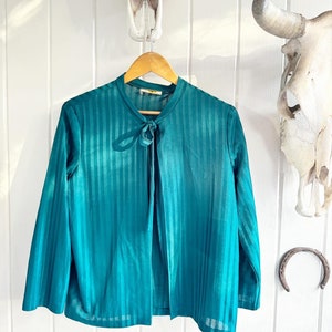 May include: A teal blue striped long-sleeved blouse with a tie at the neck. The blouse is hanging on a wooden hanger against a white wood background.