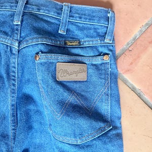 May include: Close-up of blue denim jeans with a back pocket featuring a brown leather patch with the brand name "Wrangler". The jeans have a classic five-pocket design, with copper rivets and a small brand label above the back pocket.