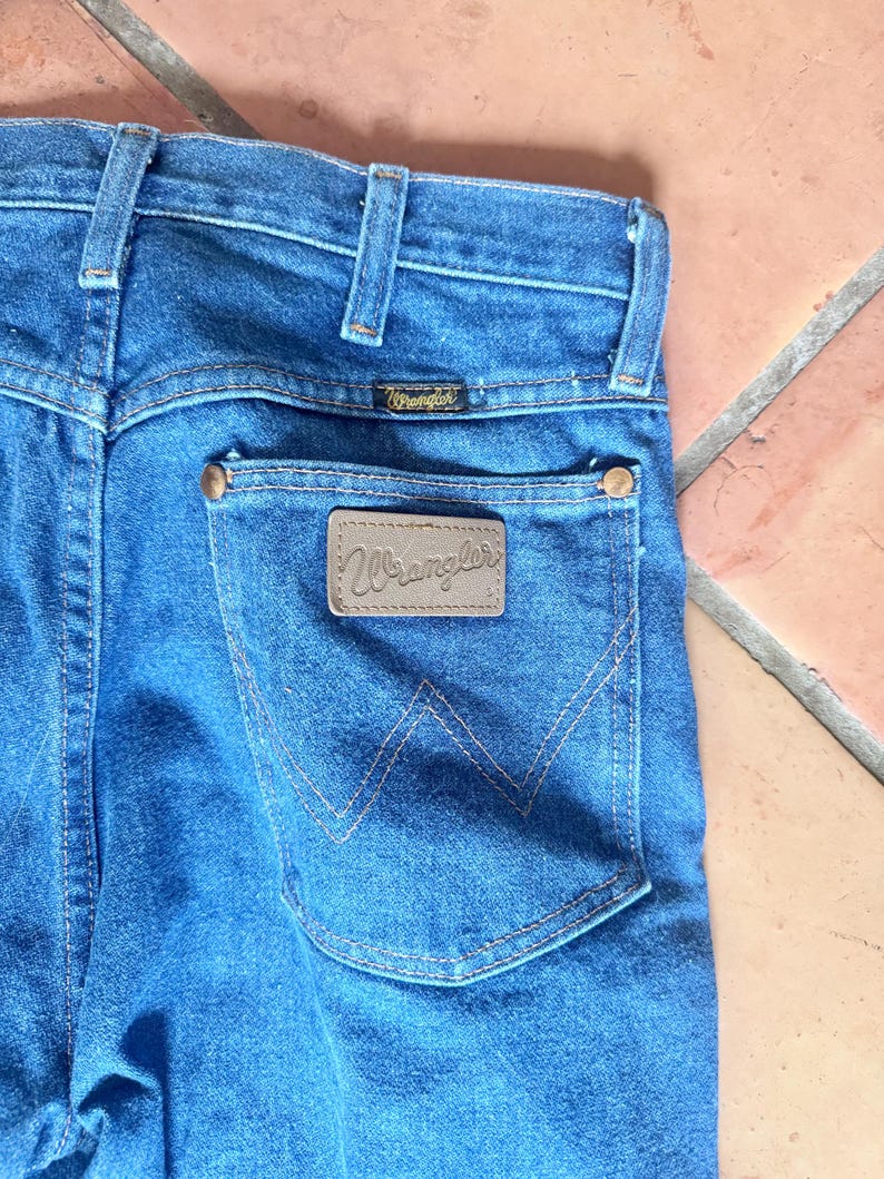 May include: Close-up of blue denim jeans with a back pocket featuring a brown leather patch with the brand name "Wrangler". The jeans have a classic five-pocket design, with copper rivets and a small brand label above the back pocket.