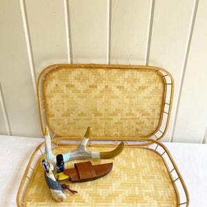 May include: Two woven bamboo trays with a natural finish. The top tray is empty and the bottom tray has a small bottle of essential oil, a leather knife sheath, and a pair of painted antlers.