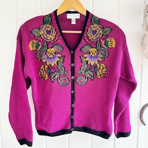 May include: A magenta cardigan with a black trim and floral embroidery. The embroidery features yellow, green, purple, and brown flowers.