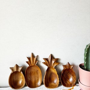 May include: Four wooden pineapple-shaped bowls, two are larger and two are smaller, all with a natural wood finish. The bowls are arranged in a row on a white surface with a white tablecloth and a pink pot with a cactus plant in the background.