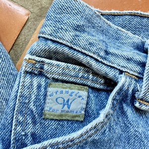 May include: Close-up of blue denim jeans with a visible pocket and waistband. The jeans feature a small rectangular tag with the brand name "Wrangler" and the letter "W" in blue. The jeans are made of a light blue denim fabric.