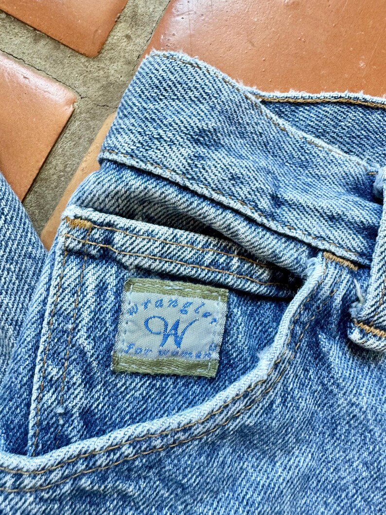 May include: Close-up of blue denim jeans with a visible pocket and waistband. The jeans feature a small rectangular tag with the brand name "Wrangler" and the letter "W" in blue. The jeans are made of a light blue denim fabric.