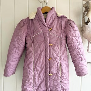May include: A purple quilted jacket with a white trim and wooden button closures. The jacket has a hood and a drawstring at the waist.