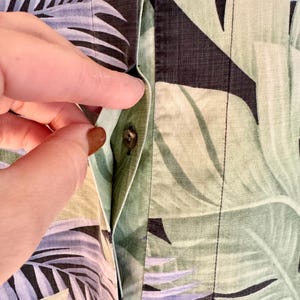 Peut inclure: Gros plan sur une chemise boutonn&eacute;e avec un motif de feuilles tropicales dans des tons de vert, noir et bleu. La poche de la chemise est visible et une main atteint le bouton.