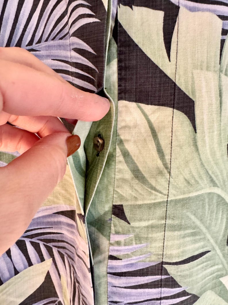 Peut inclure: Gros plan sur une chemise boutonn&eacute;e avec un motif de feuilles tropicales dans des tons de vert, noir et bleu. La poche de la chemise est visible et une main atteint le bouton.