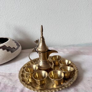 May include: A vintage brass coffee set, including a tall, ornate coffee pot with a curved spout and six small cups, all resting on a round, scalloped tray. The set is made of shiny brass and is likely used for serving coffee or tea.