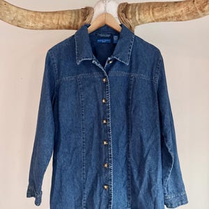 May include: A blue denim button-up shirt with long sleeves and a collared neckline. The shirt has gold-colored buttons and is hanging on a wooden hanger. The brand name "Baker Scott" is visible on the label.