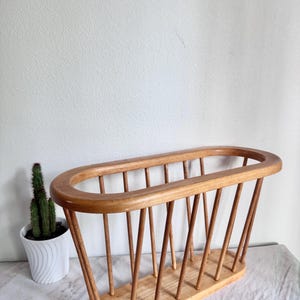 May include: A light brown wooden magazine rack featuring an oval-shaped top rim and a matching oval base, connected by a series of vertical wooden dowels that create an open, slatted structure for holding reading materials or documents.