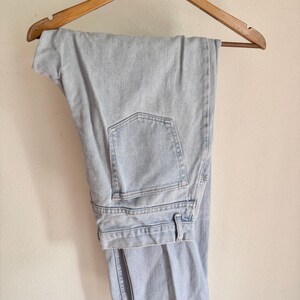 May include: Light blue denim jeans hanging on a wooden hanger. The jeans have a classic five-pocket design and a straight leg cut. The denim appears to be a light wash, with visible stitching details.