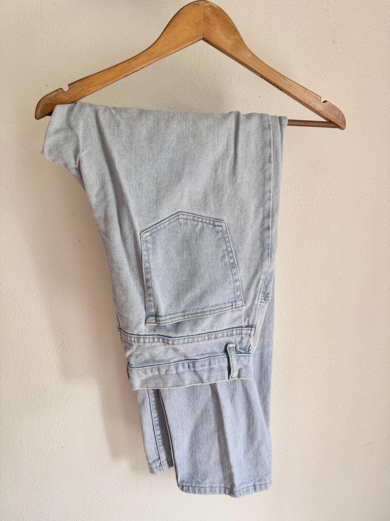 May include: Light blue denim jeans hanging on a wooden hanger. The jeans have a classic five-pocket design and a straight leg cut. The denim appears to be a light wash, with visible stitching details.