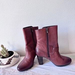 May include: A pair of burgundy leather ankle boots with side zippers and stacked heels. The boots have a classic design with a buckle detail on the side. The boots are displayed on a patterned surface.