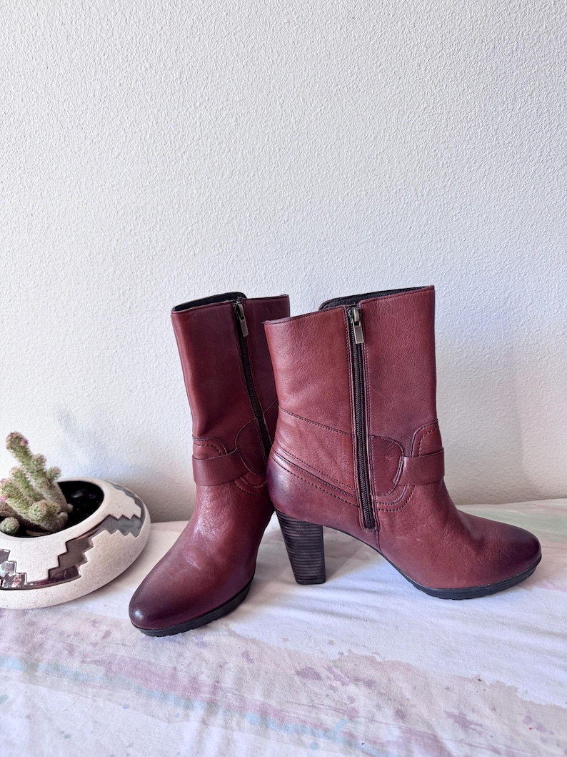 May include: A pair of burgundy leather ankle boots with side zippers and stacked heels. The boots have a classic design with a buckle detail on the side. The boots are displayed on a patterned surface.