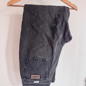 Op de afbeelding: Donkergrijze spijkerbroek die aan een houten kleerhanger hangt. De jeans heeft een klassiek ontwerp met vijf zakken en een bruine leren patch op de achterzak. Het Wrangler-logo is zichtbaar op de achterzak.