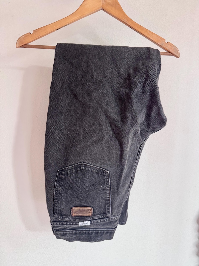 Op de afbeelding: Donkergrijze spijkerbroek die aan een houten kleerhanger hangt. De jeans heeft een klassiek ontwerp met vijf zakken en een bruine leren patch op de achterzak. Het Wrangler-logo is zichtbaar op de achterzak.