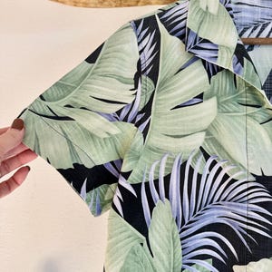 Peut inclure: Une chemise &agrave; manches courtes avec un imprim&eacute; de feuilles tropicales. La chemise pr&eacute;sente de grandes feuilles vertes avec des accents bleus et blancs sur un fond noir. Le col et la manche sont visibles.